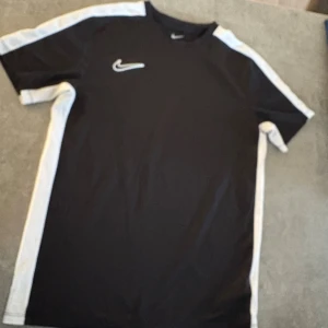 Nike Dri-Fit svart t-shirt XL - Svart Nike Dri-Fit t-shirt i funktionsmaterial med vita paneler längs sidorna och axlarna. Klassisk rund hals och korta ärmar. Perfekt för dig som gillar sportig stil och vill ha något som andas under träning.