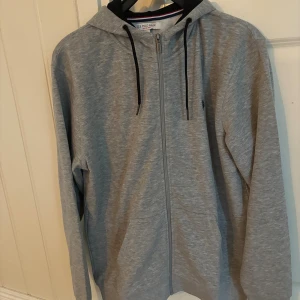 Grå hoodie från U.S. Polo Assn. - Säljer en stilren grå hoodie från U.S. Polo Assn. med dragkedja och svarta snören i huvan. Tröjan har en klassisk passform, långa ärmar och är tillverkad i mjukt bomullsmaterial. Perfekt för chill dagar eller när du vill ha en enkel look.