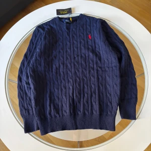 Marinblå kabelstickad tröja Polo Ralph Lauren - Kabelstickad marinblå tröja från Polo Ralph Lauren med klassisk rund halsringning och röd broderad logga på bröstet. Tröjan är i mjuk bomull och har ribbade muddar vid ärmslut och nederkant. Perfekt för dig som gillar stilren och tidlös design.