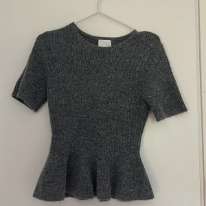 Grå stickad topp från H&M - Säljer en grå stickad topp från H&M i storlek S. Toppen har korta ärmar, rund hals och en peplumformad nederdel som ger en snygg siluett. Perfekt för dig som gillar mjuka material och enkel stil med en twist.