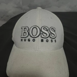 Vit keps från Hugo Boss - Snygg vit keps från Hugo Boss med stor broderad BOSS-logga framtill och justerbart metallspänne bak. Kepsen är tillverkad i bomull och har en klassisk form med böjd skärm och ventilerande hål. Perfekt för dig som vill ha en clean och stilren look.