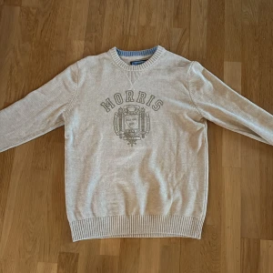 Beige stickad tröja från Morris Co - Snygg beige stickad tröja från Morris Co med rund hals och ribbade muddar. Framsidan har ett stort tryck med Morris-logga och emblem. Perfekt för dig som gillar klassisk och stilren design.