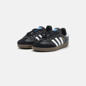 Adidas Samba svarta sneakers - Adidas Samba sneakers i svart skinn med klassiska vita ränder på sidorna och brun gummisula. Skorna har snörning, rund tå och låg profil. Ikonisk blå Adidas-logga på plösen och gul Samba-text på sidan. Perfekt för dig som gillar retrostil och streetwear.Inha defekter vad jag kan se, bara lite sliten på sulan, se bild 💕