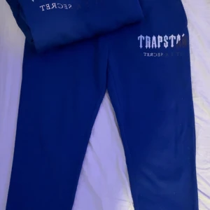Blå Trapstar tracksuit med tryck - Snygg blå hoodie från Trapstar med stort vitt tryck på bröstet och snörning i huvan. Tröjan har en klassisk känguruficka framtill och är gjord i mjukt bomullsmaterial. Hoodien och byxorna är i storlek L men passar också M