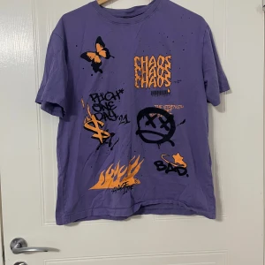 Lila t-shirt med tryck från Black Squad - Lila t-shirt från Black Squad med coola tryck i orange och svart, bland annat fjärilar, eldflammor, panter och graffiti-text. Modellen är relaxed fit och har korta ärmar. Tillverkad i 100% bomull för en skön känsla.