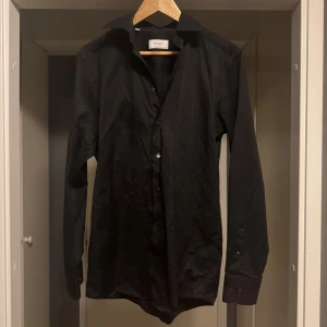Svart skjorta från Eton - Klassisk svart skjorta från Eton med lång ärm och knappar framtill. Skjortan har en stilren design med bred krage och är tillverkad i ett slätt bomullstyg. Perfekt för dig som gillar en clean och tidlös look.
