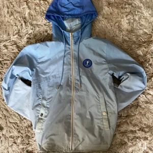 Blå vindjacka från Trapstar med huva - Snygg blå vindjacka från Trapstar med huva och dragkedja framtill. Jackan har broderad Trapstar-logga på ryggen och ett blått märke på bröstet. Justerbar huva med snörning och praktiska fickor med knappar. Perfekt för dig som gillar streetwear vibes. priset är ej hugget och svarar gärna på frågor