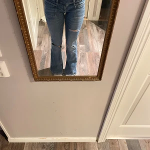 Gina Tricot jeans med slitningar - Säljer ett par blå bootcut jeans med låg midja och slitna detaljer vid knäna. Jeansen har framfickor och klassisk jeanslook, perfekta för en avslappnad och trendig stil. Materialet är jeans och passformen är normal.