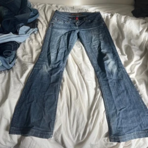 Blå bootcut jeans med låg midja - Snygga blå jeans från Hilfiger Denim (river island) med bootcut passform och låg midja. Klassisk femficksmodell med slitningar och ljusare partier på låren. Jeansen har dragkedja och knapp framtill samt Hilfiger-detaljer på fickorna. En liten defekt nedtill men det är inget man tänker på! innerbenslängden är ca 76cm skriv vid frågor🫶