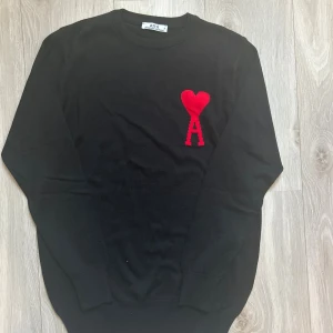 Svart tröja från Ami Paris - Svart tröja från Ami Paris med röd broderad logga i form av ett hjärta och bokstaven A på bröstet. Tröjan har rund halsringning och långa ärmar, perfekt för dig som gillar stilrena och trendiga plagg. Skriv privat för mer frågor 