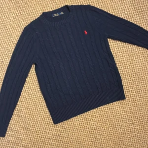 Mörkblå kabelstickad tröja Polo Ralph Lauren - Kabelstickad mörkblå tröja från Polo Ralph Lauren med klassisk rund halsringning och röd broderad logga på bröstet. Tröjan har långa ärmar och ribbade muddar för en snygg passform. Perfekt för dig som gillar stilren och tidlös design. Storlek L men passar mer som M.