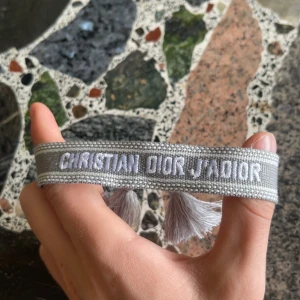 Grå Christian Dior J'Adior armband - Snyggt grått armband från Christian Dior med broderad text 'J'Adior' i vitt. Armbandet har dekorativa tofsar i ändarna och är gjort i ett mjukt tygmaterial. Perfekt för dig som vill ha en lyxig och trendig accessoar med designer-vibe.