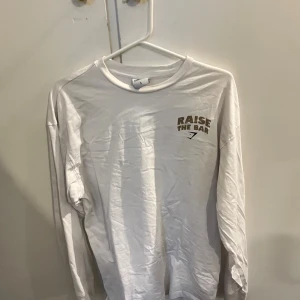 Vit långärmad Gymshark-tröja S - Vit långärmad tröja från Gymshark med trycket 'RAISE THE BAR' i guld och svart framtill och stort motiv på ryggen. Perfekt för dig som gillar träning och vill sticka ut på gymmet. Mjuk bomullskänsla och relaxed fit.Passar M också 