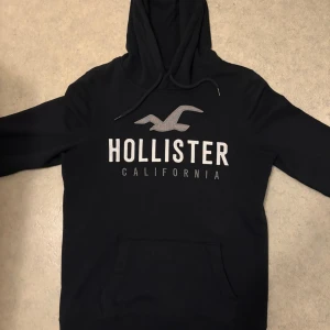 Svart hoodie från Hollister - Svart hoodie från Hollister med stor logga och text på bröstet. Klassisk känguruficka framtill, huva med snörning och mjukt material. Perfekt för chill dagar eller när du vill ha en enkel och snygg look. Otrolig passform.