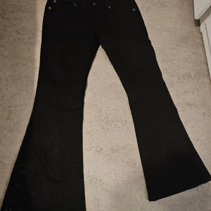 Svarta bootcut jeans från Fishbone - Svarta jeans från Fishbone i klassisk bootcut-modell. Byxorna har fem fickor, silverfärgade nitar och knapp, samt Fishbone-logga på bakfickan. Snygg passform med utsvängda ben och midjehög skärning. Perfekta för dig som gillar en retrostil.