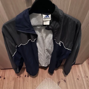 Adidas vintage vindjacka blå/grå - Retro vindjacka från Adidas i blått och mörkgrått med vit piping längs sömmarna. Jackan har hög krage, elastiska muddar och klassisk sportig look. Perfekt för dig som gillar oldschool streetstyle och sportmode.