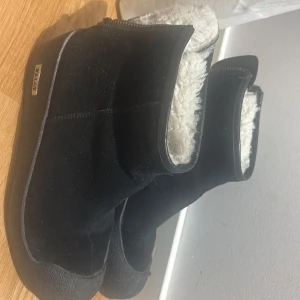 Svarta mockaboots med dragkedja( Bally ) - Svarta boots i mocka från EMU Australia med mjukt vitt foder och dragkedja baktill. Skorna har en rund tå och platt sula, perfekta för kalla dagar. Insidan är fodrad med fluffigt material för extra värme och komfort.