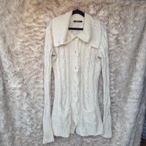 Vit stickad kofta från Gina Tricot - Lång vit kofta med stora knappar. Koftan har en stor krage. Länga muddar vid ärmsluten. Fickor fram. Knapparna är stora så hålen har blivit lite uttöjda. Fint stickat mönster.