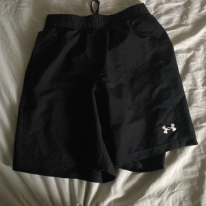 Svarta träningsshorts Under Armour og pris 799 - Svarta träningsshorts från Under Armour med elastisk midja och vit logga på benet. Shortsen har en loose passform och är gjorda i lätt polyester som andas. Midjan har en bred resår med Under Armour-tryck i vitt för extra sportig vibe.