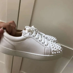 Vita Louboutin sneakers med nitar - Säljer ett par vita sneakers från Christian Louboutin med ikonisk röd sula och coola nitar över tån. Skorna har snörning och är tillverkade i skinn, vilket ger en lyxig känsla. Perfekta för dig som vill sticka ut med en edgy och trendig stil.