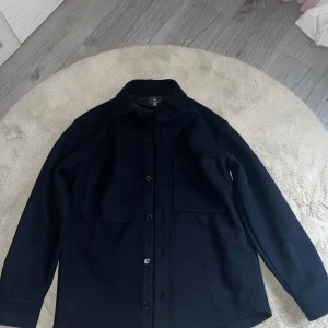 Mörkblå overshirt från H&M - Snygg mörkblå overshirt från H&M med relaxed fit. Skjortan har två stora bröstfickor, knäppning framtill och klassisk krage. Perfekt lager-på-lager-plagg med lång ärm och stilren look. Materialet är ullblandning som ger en mjuk och varm känsla. 