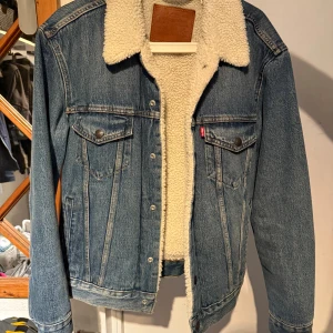 Levi's jeansjacka med teddyfoder S - Klassisk blå jeansjacka från Levi's med vitt teddyfoder och krage. Jackan har två bröstfickor med knappar, knappstängning framtill och detaljer i silver. Perfekt för kyliga dagar och ger en snygg vintagekänsla.