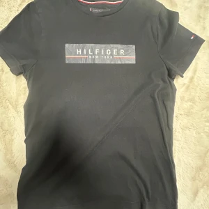 Svart t-shirt Tommy Hilfiger - Svart t-shirt från Tommy Hilfiger med tryckt logga 'Hilfiger New York' på bröstet. Klassisk rund halsringning och korta ärmar. Skön passform och diskret liten flagga på ärmen. Perfekt för en stilren och enkel look.