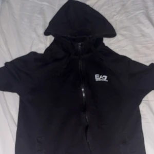 Svart hoodie från EA7 Emporio Armani - Svart hoodie från EA7 Emporio Armani med dragkedja framtill och huva med snörning. Tröjan har en stilren look med EA7-logga på bröstet och praktiska fickor. Perfekt för dig som gillar streetwear och vill ha något bekvämt och snyggt.