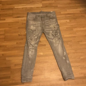 Grå slitna jeans från Dsquared2 - Säljer ett par grå jeans från Dsquared2 med coola slitningar och färgstänk. Jeansen har skinny passform och klassisk femficksdesign. Perfekta för dig som gillar en edgy streetstil. Materialet är jeans och färgen är ljusgrå. 