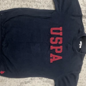 Mörkblå tröja från US Polo Assn - Mörkblå tröja från US Polo Assn med stora röda bokstäver 'USPA' på bröstet och broderad logga nertill. Klassisk rund halsringning och ribbade muddar. Perfekt för dig som gillar sportig och stilren look.