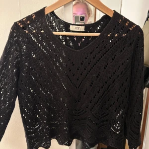 Svart stickad croppad tröja - Svart stickad tröja med hålmönster och croppad passform. Tröjan har trekvartsärmar och en snygg vågig kant nertill. Perfekt att styla med jeans eller kjol för en cool look.