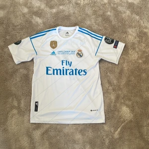 Vit Real Madrid fotbollströja - Säljer en vit Real Madrid fotbollströja från Adidas med blå detaljer och text. Tröjan har korta ärmar och tryck med 'Ronaldo' och nummer 7 på ryggen. Champions League-finalen i Cardiff 2017 är tryckt på framsidan. Perfekt för fans av laget och spelaren!