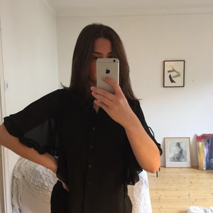 Blus med volang - En svart blus i tunt, lite genomskinligt tyg. Blusen har fladermusarmar med volang som faller fint samt knäppning hela vägen upp. Aldrig använt.