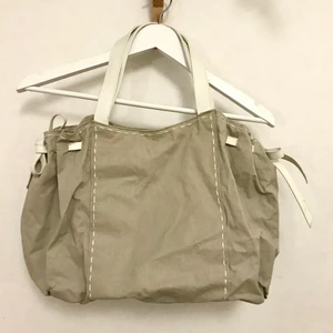 Beige väska, vita handtag och band  - En ljust beige väska i litet stelare tyg med vita ”skinn”-handtag och band som går att dra åt och knyta i sidorna. Helt oanvänd! Jättefint skick. Från Lancôme. Ca. 30x40 cm. Kan mötas i Stockholm eller skicka mot fraktkostnad! ✨🌸✨