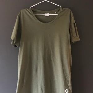Mörkgrön t-shirt - Säljer mörkgrön t-shirt, aldrig använd. En detalj är att det sitter en dragkedja på vänsterarmen.
