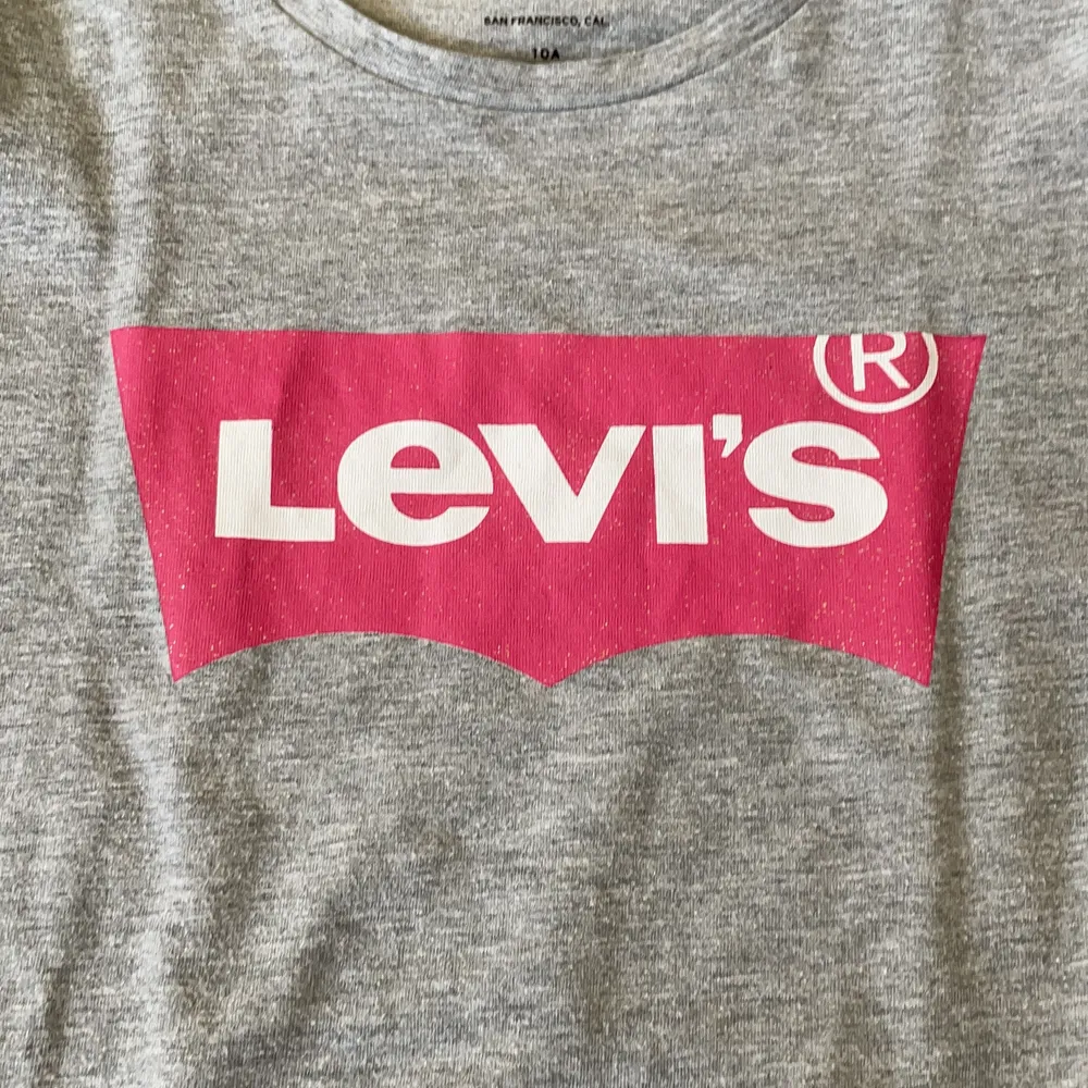 En grå tröja med ett tryck med märket Levis och med rosa färg!. T-shirts.