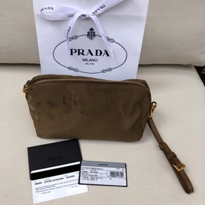 PRADA väska/sminkväska/necessär - PRADA nylon jacquard contenitore maniglia i militärgrönt/brun med detaljer i guld samt brunt läder. Insidan har det klassiska PRADA-trycket samt en ficka med dragkedja. Både utsida och insida är lätta att torka av.   Går att använda både som necessär, sminkväska eller som en liten handväska/pouch/clutch. Har ett läderband som går att ta bort vilket gör att den kan användas som väska och bäras runt handleden (som en wristlet).  I nyskick och finns inga tecken på användning. Fler bilder kan såklart skickas. Kommer med kvitto, äkthetsbevis, påse samt prada-band.  MÅTT: Längd 19,5cm  Höjd 12cm Djup 9cm! Men den är ju i mjukt material så den är lätt att ”fylla upp”!