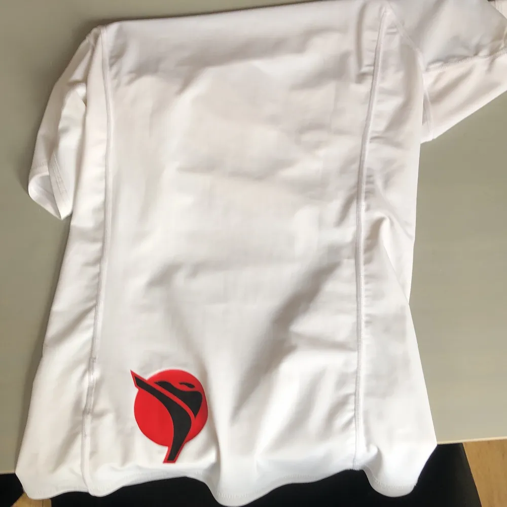 Jättefin vit tight T-shirt i träningdmaterial med liten polokrage. På ena ärmen står det ”budo nord” i svarta bokstäver, och på ryggen finns ett röd/svart kampsportsmärke. Jätteskön och ej genomskinlig💕. T-shirts.