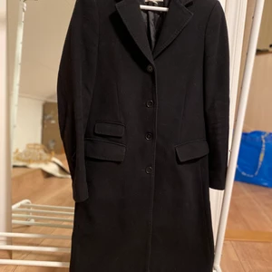 Kappa/Rock - Figursydd klassisk kappa i 75% ull 20%polyamid 5%cashmere            Svart kappa, två fickor fram, enkel knäppning. Baktill sprund            Ca knälång på 165cm lång person.         NY pris 5.600kr 