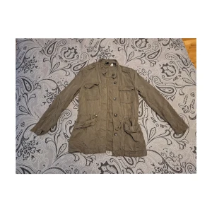 Vår/höstjacka - En tunn militärgrön jacka med resårband i midjan, 4 fickor där fram och knappar. Väldigt skön och snygg att styla sin outfit med när det börjar bli lite svalare ute