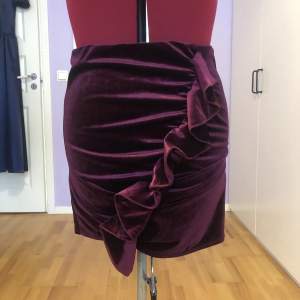 plommon-färgad kjol i sammet liknande tyg med volang som går på ena sidan kjolen provdocka mått:  midja: 69 cm höft: 91 cm