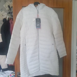 Vit täckkappa, tuxer, strl xl - En offwhite, aldrig använd täckkappa från tuxer i storlek xl. Den har en ej avtagbar luva, tvåvägsdragkedja och två fickor fram, med knapp.