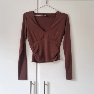 Långärmad topp - En brun långärmad topp köpt här på plick. Den har djup ringning och rynkade detaljer i sidorna. Någon tråd lös längs med linningen men inget som stör.  Material: Yttertyg 93% återvunnen polyester 7% elastan Innertyg 100% polyester