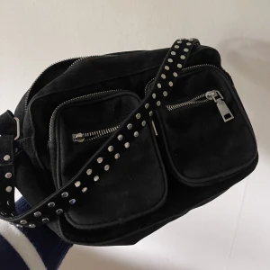 Svart handväska - Mockaväska från Noella. Modellen heter Celina. Det är en liten väska men mycket får plats i den och det finns även små fack både i väskan och utanpå. Finns ett längre axelband som medföljer väskan. Saknas en bit av dragkedjan på vänster sida (se bild)