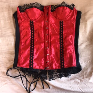 Korsett - En röd och svart korsett som jag har köpt second hand. Har endast provat den(den var för stor). Det finns några hål i spetsen och en lös söm där bak. Den är lite böjd också (se bild 1). Den är i storlek 36!