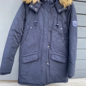 Dunparkas Svea - Superfin dunjacka från SVEA, storlek 170. Marinblå, med SVEA broderat ton-i-ton på bröstet och SVEA-märke på höger ärm. Luva med avtagbar pälskant. Parkasmodell. Sparsamt använd, som ny. Köparen betalar frakt, eller hämtar hos mig.