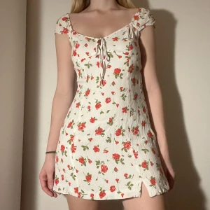 Vit gullig klänning med röda blommor - Söt vit sommarklänning med mönster av röda rosor och gröna blad. Den har ett band som knyts där uppe. Ganska kort på mig som är 165 cm. Den har dragkedja på sidan så lätt att ta på sig. Står storlek M men snarare xs. Kan mötas nära Mariatorget eller posta