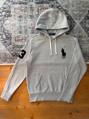 Grå Polo Ralph Lauren Hoodie - Big Pony - Hej! 👋 Säljer en riktigt eftertraktad grå Polo Ralph Lauren ”Big Pony” hoodie i storlek M. Har den stora broderade svarta loggan på bröstet och siffran ”3” på höger ärm. 🐎✨ Tröjan är i mycket gott skick, välvårdad och helt utan hål eller fläckar. Ett perfekt premium-basplagg som passar till exakt allt i garderoben och håller stilen året om! Garanterat äkta med tvättlappar kvar. Fraktar snabbt! 💯 🚛📦 