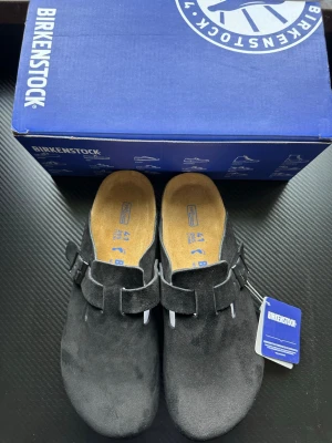 Svarta Birkenstock tofflor i mocka - Säljer ett par svarta Birkenstock tofflor i mjuk mocka med justerbart spänne och korksula. Klassisk slip-in modell med bekväm fotbädd och diskret logga på sidan. Perfekta för chill dagar hemma eller när du vill ha det extra bekvämt.