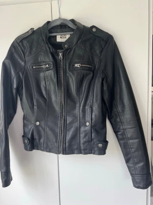 Svart ”skinnjacka” (ej äkta) från Vero Moda - Snygg svart ”skinnjacka” (ej äkta) från Vero Moda med dragkedja framtill, två bröstfickor med dragkedja och dekorativa axelknappar. Jackan har figurnära passform och quiltade detaljer på axlarna. Perfekt för en cool och trendig look.
